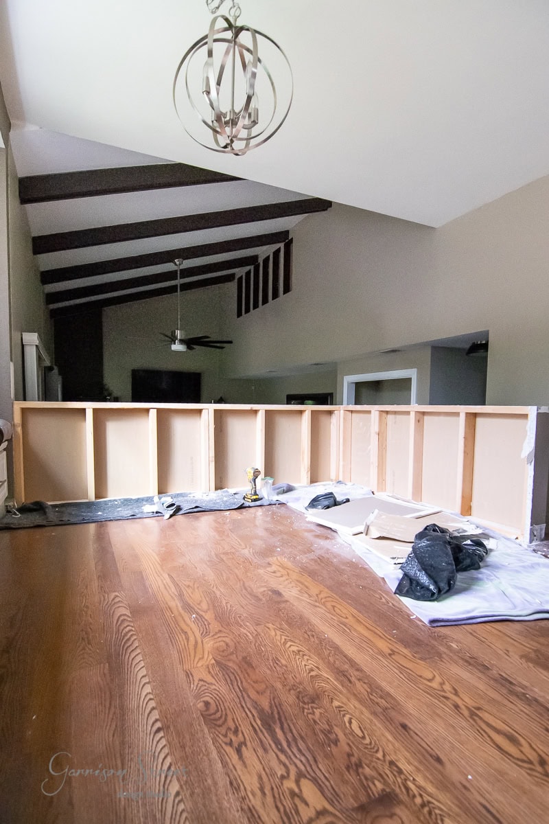 A home interior under renovation showcases an unfinished kitchen counter with exposed cabinets, surrounded by brown hardwood flooring. A tarp covers part of the floor. The room, featuring vaulted ceilings and visible support beams, embodies a DIY spirit with a light fixture awaiting installation.