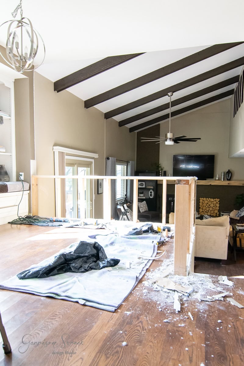 A living room with high ceilings and exposed beams is under renovation, with plans to install a sleek cable railing. Tarps cover the hardwood floor, and construction debris is scattered around. A large window lets in natural light, and a ceiling fan is visible above.
