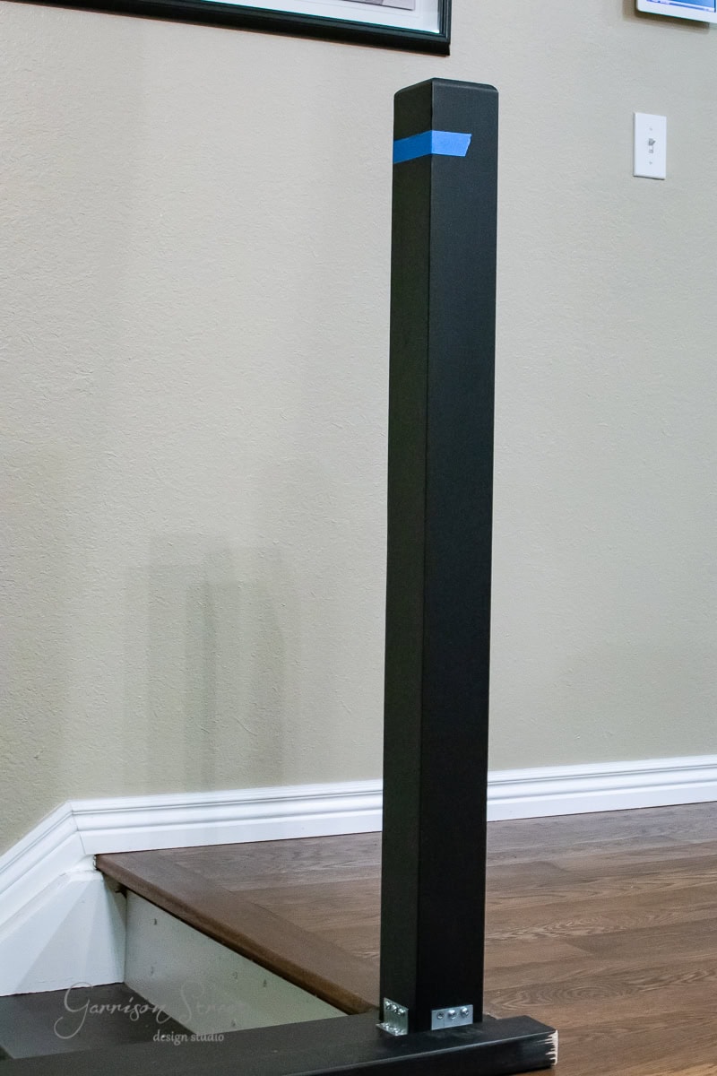 A black metal post stands at the corner of the wooden staircase, secured with metal brackets. Blue tape near the top marks its installation. The staircase railing leads to a beige wall where a white electrical outlet is visible, perfect for your next DIY touch or cable management solution.