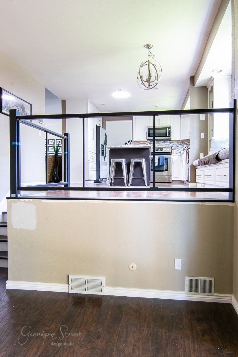 A modern interior boasts a split-level open floor plan. The raised kitchen area, with counter and stools, is separated by a sleek cable railing. The lower cozy area features dark hardwood floors and soft neutral walls. Overhead, a modern chandelier casts its glow.