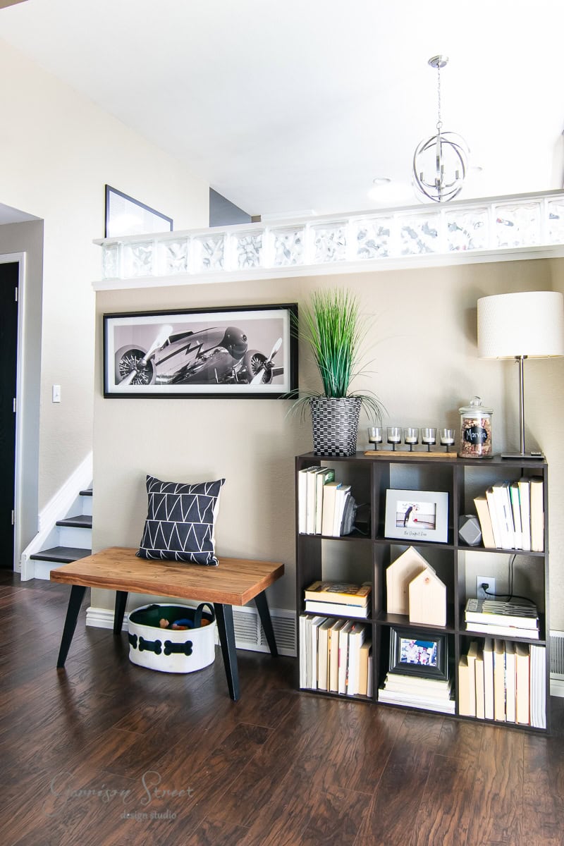 A modern, cozy living space with a dark wood floor features a casual bench with a cushion. A black shelf holds books, decor, and a plant. A framed motorcycle photo hangs above, and a tall lamp stands beside the shelf. A clear wall divider adds light to this stylish makeover.