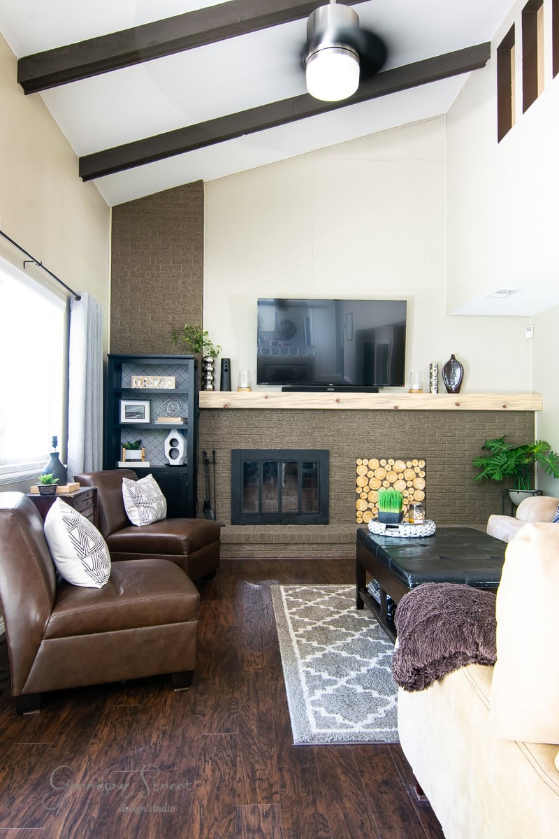 A cozy, modern living room with high ceilings and wooden beams. A wall-mounted TV is above the fireplace, complementing the brown leather chairs surrounding a dark coffee table. The beige sofa and decorative shelves add warmth to this casually elegant space, perfect for a stylish makeover.