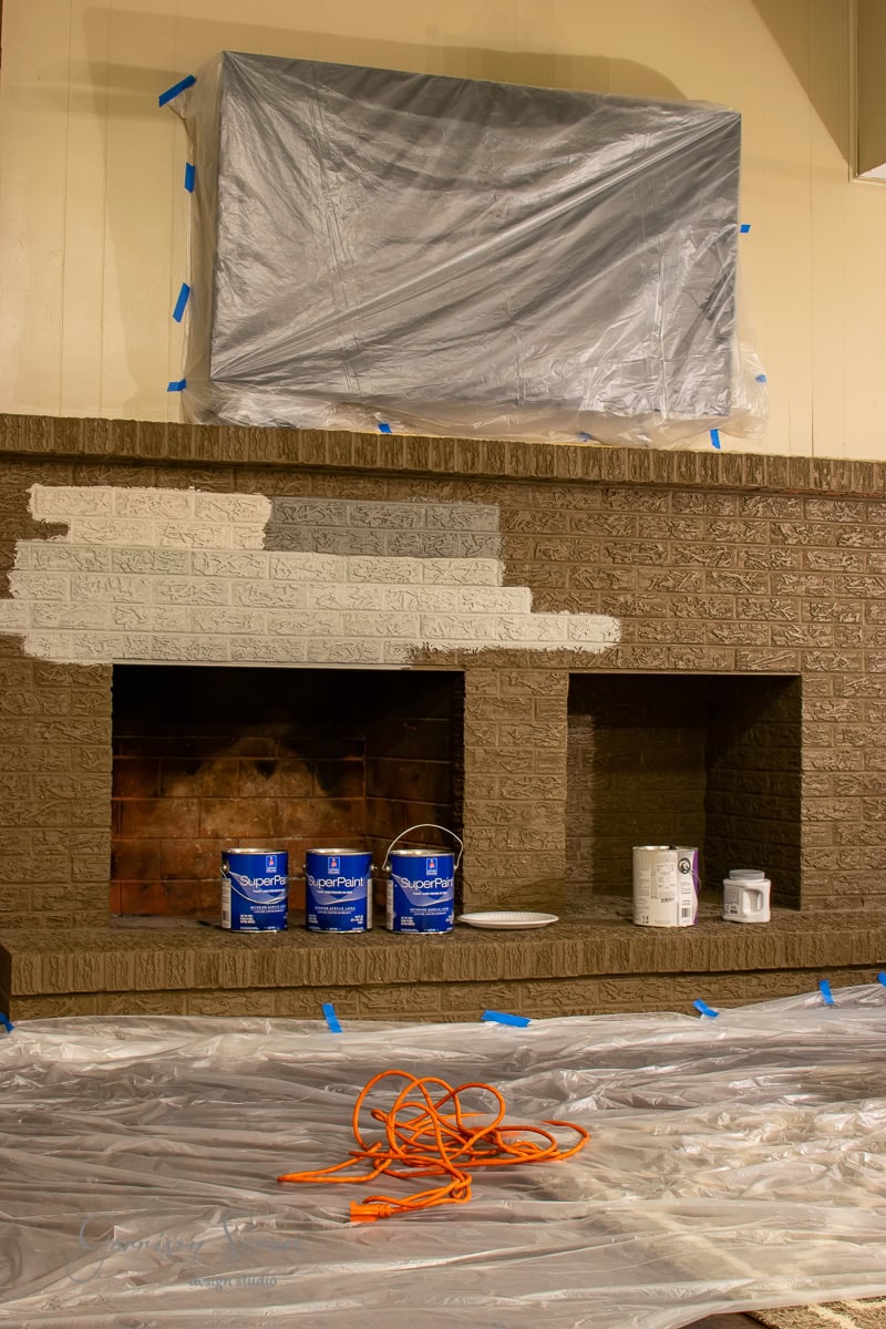A DIY brick fireplace makeover unfolds with three cans of paint resting on the hearth. A plastic sheet covers an area above the mantle, while an orange extension cord lies coiled on the floor, which is safely covered with protective plastic.