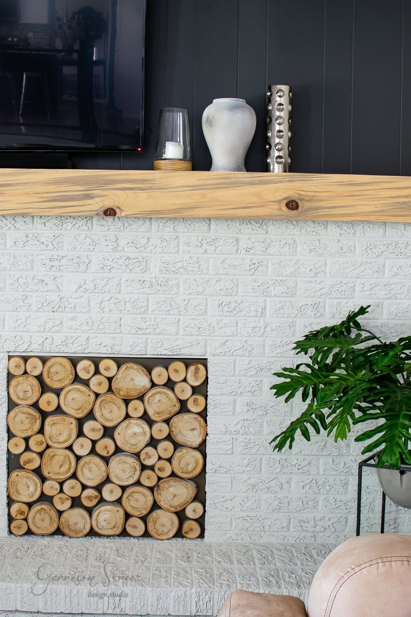 A cozy living room corner. The white brick surrounds a decorative display of cut wood logs in the opening. A wooden mantel holds a vase, candle, and metal art piece. A green plant adds life on the side, beneath the mounted TV.