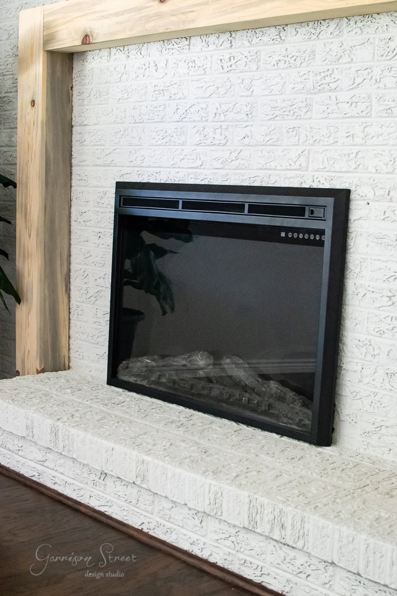 A modern electric fireplace with a black frame sits elegantly against a white painted brick wall. The light wood mantel adds a rustic touch. Dark wooden floors complement the scene, while a hint of greenery peeks in from the left.