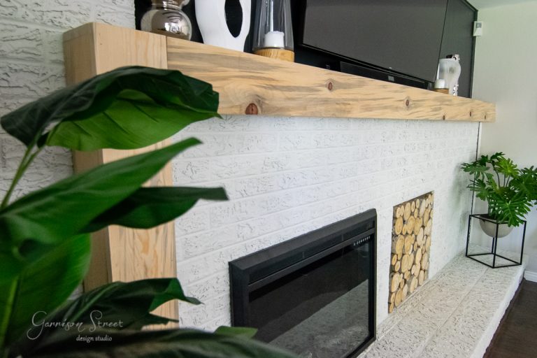 A modern living room features a white brick fireplace, showcasing a DIY makeover. The mantel displays decorative items, including a black and white vase, while a TV is mounted above. A potted plant sits beside the fireplace, which boasts a neatly stacked wood pile in its opening.