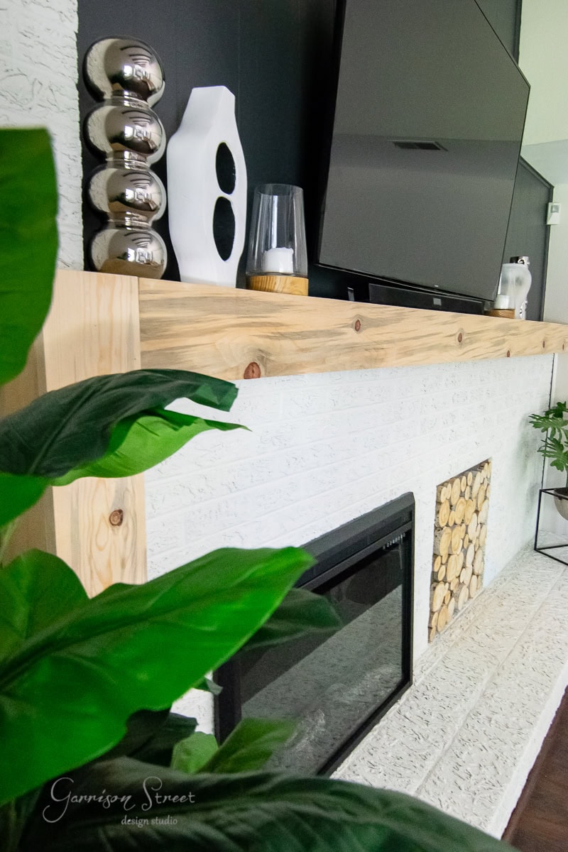 A modern living room. The white mantel displays decorative objects like silver spheres and a sleek vase. Above, a large flat-screen TV is mounted, while a vibrant green plant adds life to the space.