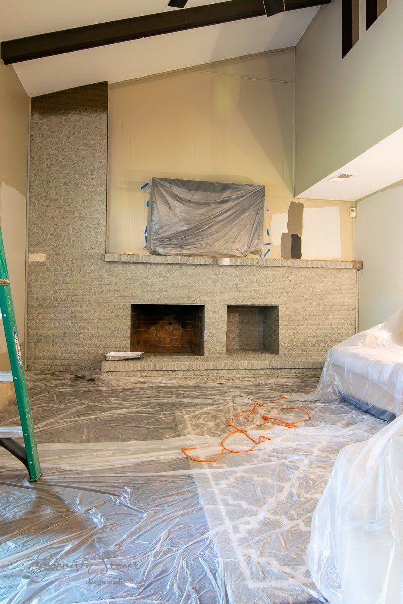 The room is undergoing a brick fireplace makeover, with the hearth currently covered in plastic sheeting. Walls are dotted with various paint swatches, and protected flooring hosts a green ladder on the left and an extension cord sprawling across it, indicating an active DIY project.
