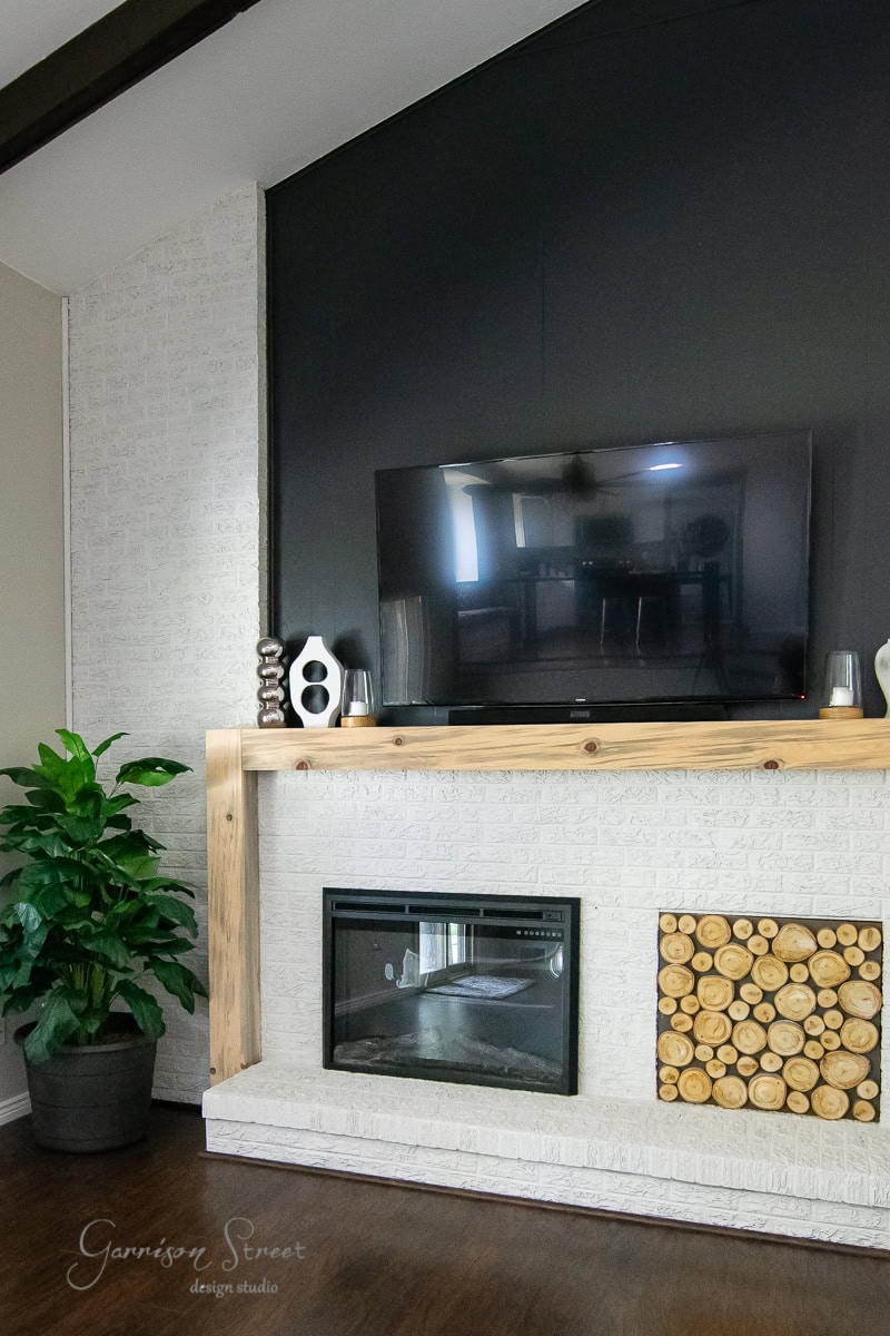 A modern living room features a black accent wall, mounted flat-screen TV, and light wooden mantel. Below, a sleek brick fireplace adds warmth with its stacked logs. A large potted plant to the left enhances the cozy atmosphere.