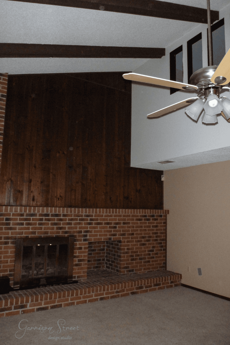 A cozy living room showcases a DIY brick fireplace makeover with a wooden mantel. The vaulted ceiling features wooden beams and a ceiling fan with lights, while the mix of wood paneling and neutral paint on the walls brings warmth to the space.