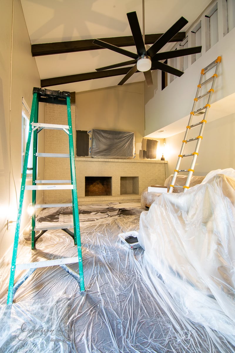 A room under renovation with high ceilings features a green ladder, a wooden one leaning against the wall, and a ceiling fan. The floor and furniture are covered with plastic sheets for a DIY brick fireplace makeover.
