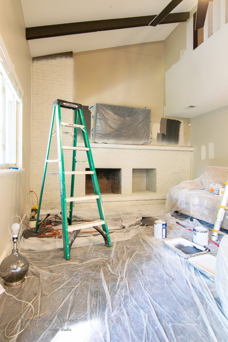 A living room under renovation features a green ladder at its center, and walls partially painted. The brick fireplace is covered with plastic, hinting at a DIY makeover. Paint cans and tools are scattered across the sheet-protected floor, alongside a lamp and a covered couch.