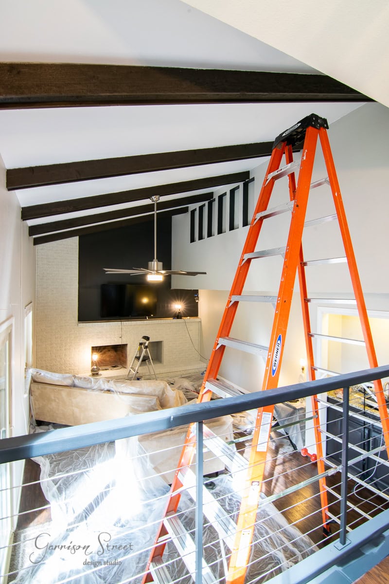 A living room with a high ceiling boasts exposed wooden beams and an inviting brick fireplace. An orange ladder is set up under the beams, hinting at a DIY makeover project. The space includes a sofa wrapped in plastic, a ceiling fan, and a flat-screen TV above the charming brick mantel.