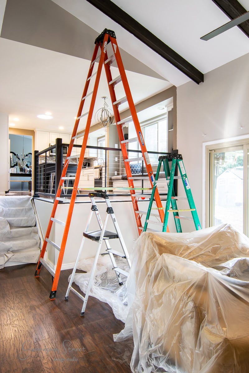 A modern room with a high ceiling features three ladders of varying sizes. The largest is red, the medium green, and the smallest silver. Amid scattered construction materials on the wooden floor, plans for a DIY brick fireplace makeover unfold as furniture remains covered with plastic sheets.