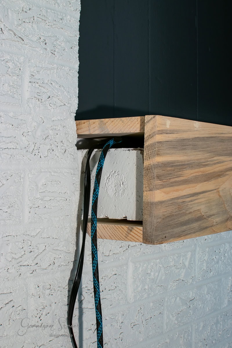 Close-up of a wooden box mounted on a white brick wall. Electrical cables and a blue-rope covered wire emerge from the base.