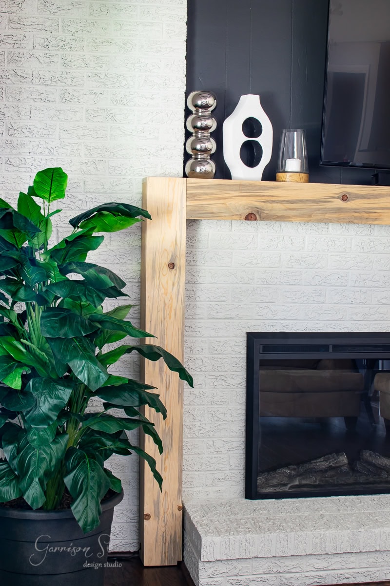 A modern living room featuring a white brick wall and wooden mantel that complement the sleek black fireplace, adorned with decorative items like a white vase. Beside it, a large green plant in a black pot adds a touch of nature's elegance.