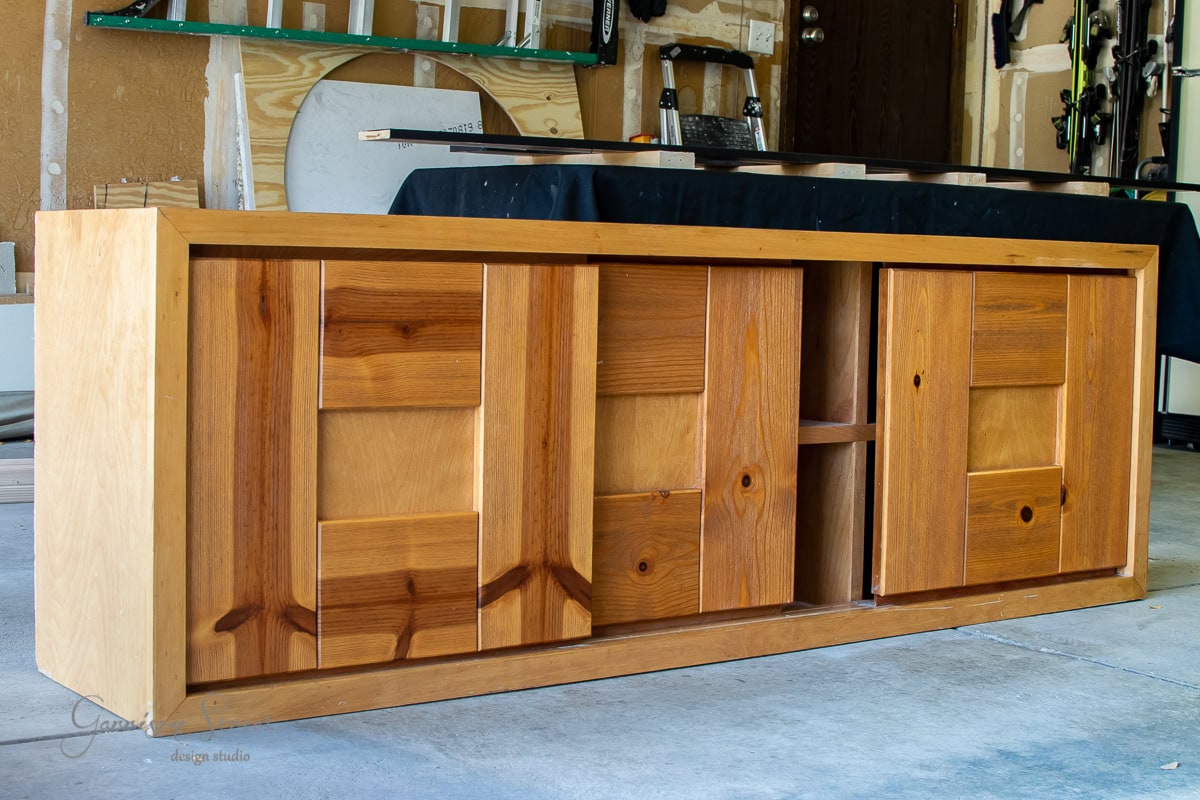 A wooden cabinet with sliding doors and a geometric pattern showcases a mid-century modern (MCM) style, perfectly complementing the concrete floor in the workshop. Various tools and materials glow up in the background, highlighting this chic piece of furniture.