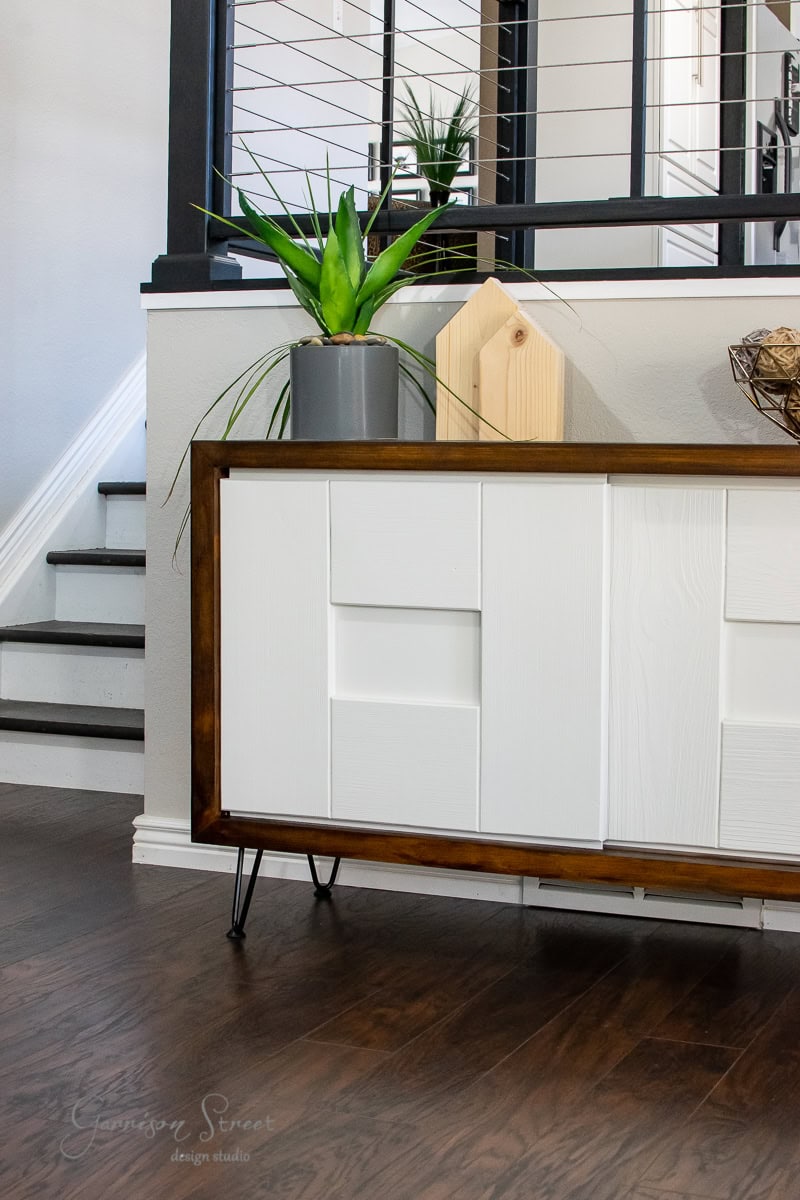 A modern MCM sideboard with a wooden frame and white panels stands on a dark wood floor, adding a stylish glow up to the space. A potted plant sits on top, accompanied by a wooden geometric decor piece. Stairs with a black metal railing are visible in the background.