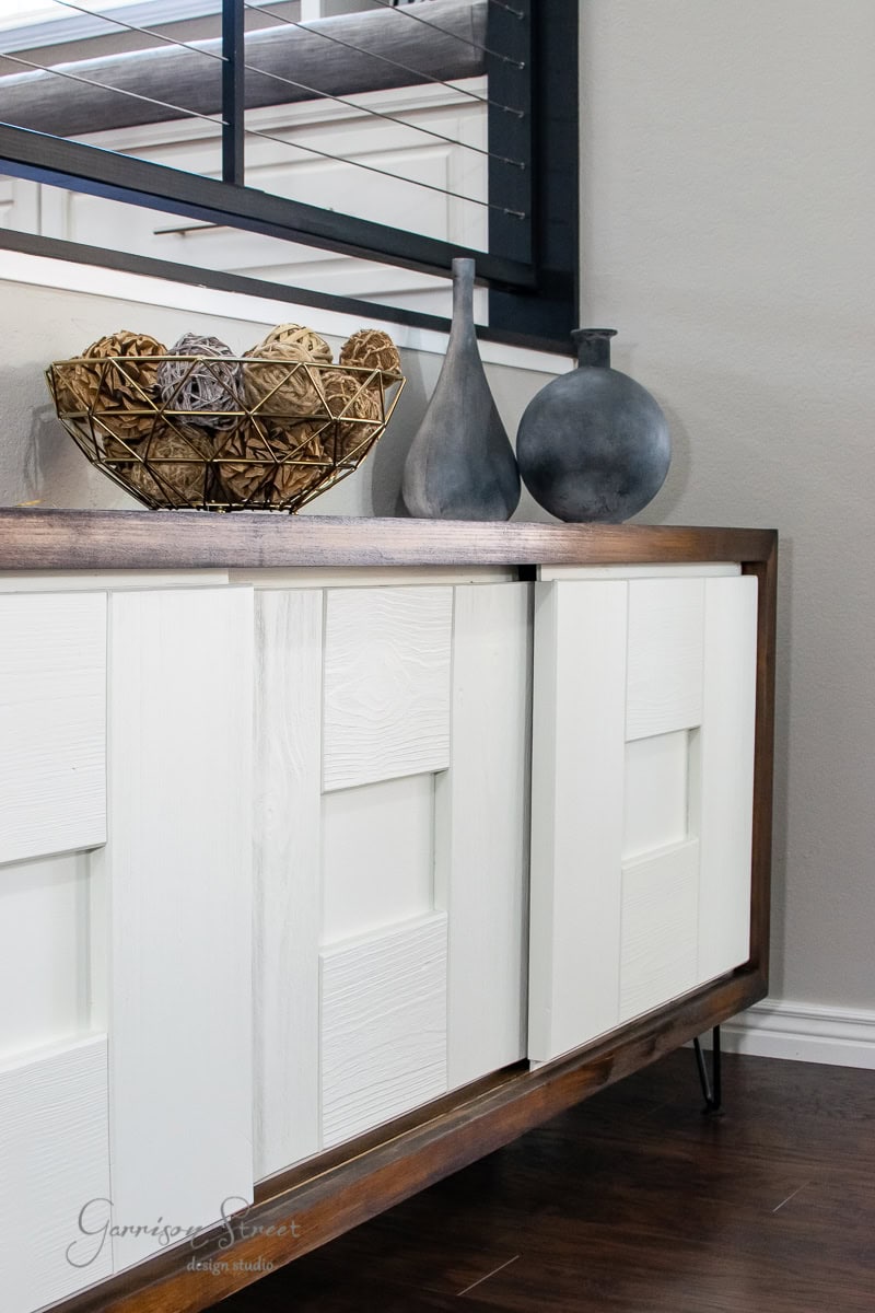 A modern sideboard with a wooden frame and white panel doors stands proudly, showcasing its MCM style. Atop, two matte gray vases and a decorative wicker basket filled with spherical ornaments complete the furniture's glow-up. A window with black frames is reflected in the mirror above.