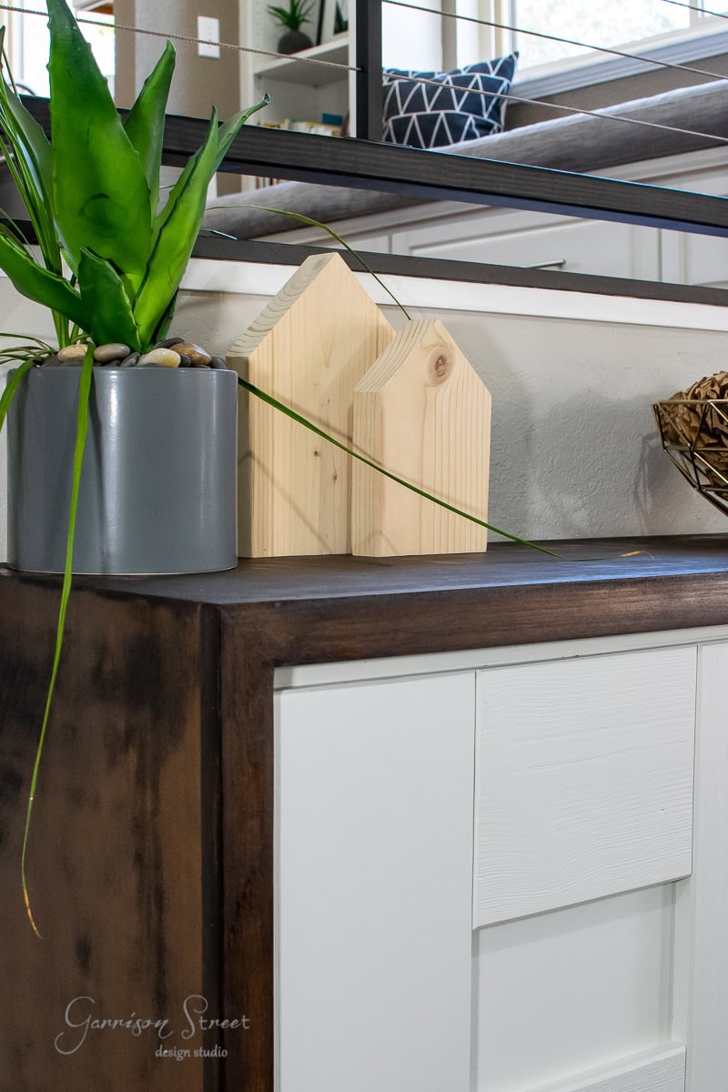 A modern MCM interior features a wooden cabinet with sleek white drawers. On top, there's a gray planter with a green plant and two wooden house-shaped decor items, giving the space a furniture glow up. A metal railing and a window are visible in the background.