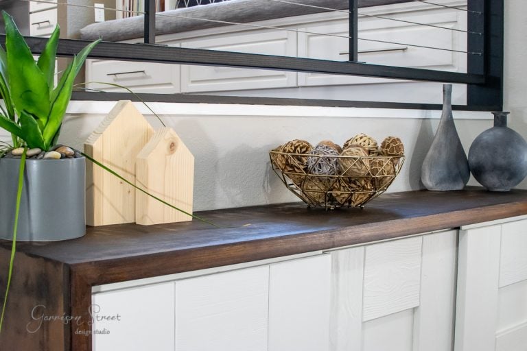 A modern interior design scene showcases an MCM-inspired wooden sideboard adorned with potted plants. Decorative wooden blocks, a wire basket filled with wicker balls, and two matte vases are elegantly placed in front of a large mirror, giving the space a stylish glow up.