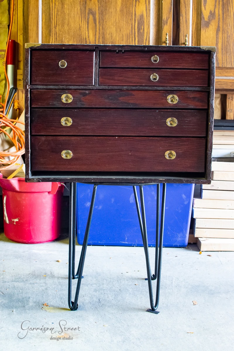 A small, dark wooden dresser with a retro MCM design boasts two top drawers and four larger lower ones, each adorned with circular brass knobs. Standing on thin metal legs, this piece adds a stylish glow up to your garage, even with storage items visible in the background.