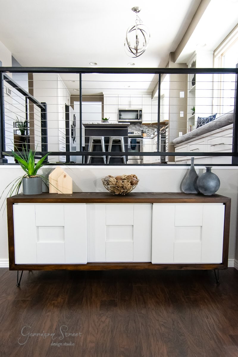 A modern room features a white and wooden credenza, showcasing a touch of MCM style, topped with vases, a plant, and decorative items. Behind it, a railing overlooks the dining area with a table and chairs. The space radiates an effortless glow-up with its minimalist design and neutral colors.