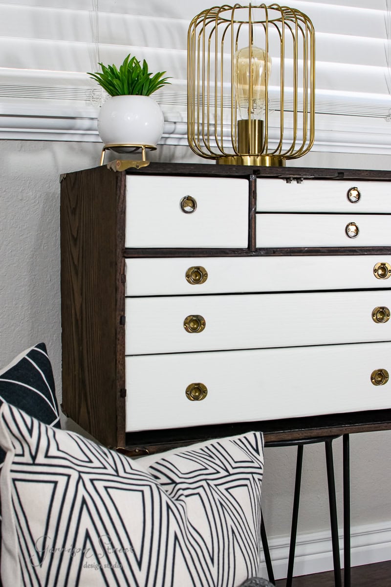 A wooden dresser with white drawers and brass knobs stands against a light gray wall, showcasing its MCM style. On top is a potted plant and a decorative lamp featuring a unique gold wire cage around the bulb. A geometric-patterned pillow lies in the foreground, completing the furniture glow up.