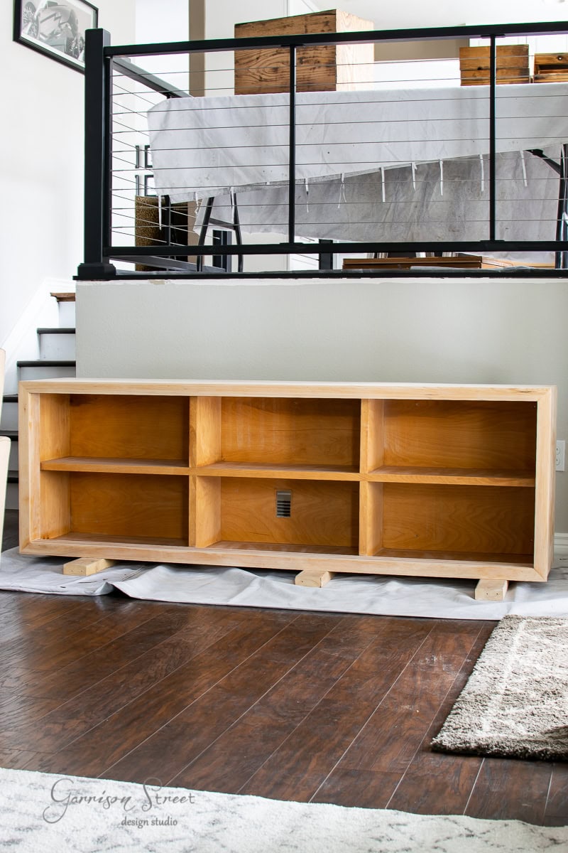 A wooden storage unit with open shelves sits on a wooden floor, exemplifying mid-century modern elegance. It’s placed in front of a black metal and glass railing, with a covered table glowing up in the background. The design studio’s name graces the bottom left.