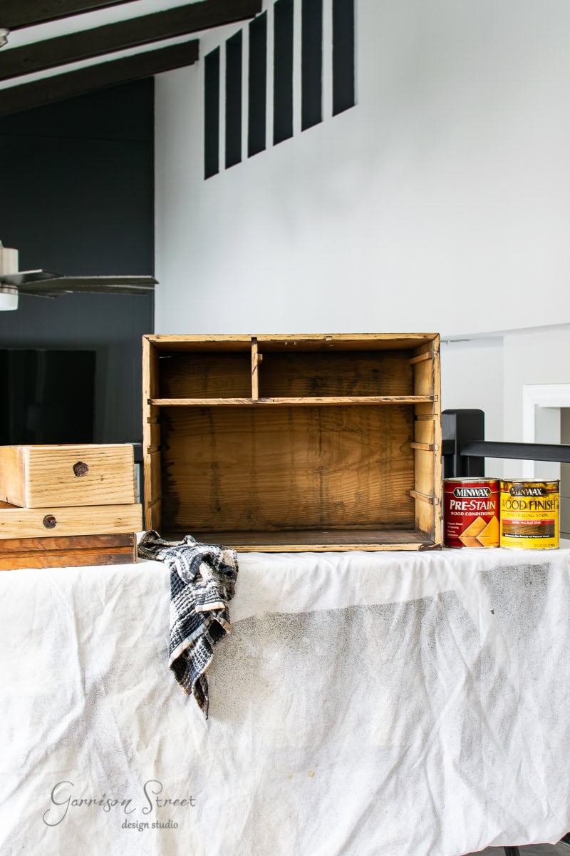 An unfinished wooden dresser sits on a covered surface, ready for its MCM glow up, surrounded by a folded towel and two tins labeled "pre-stain" and "wood finish." The background features a modern interior with black and white elements.