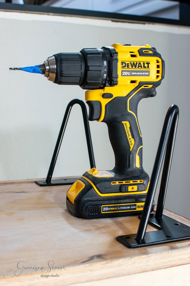 A DeWalt 20V cordless drill with a blue drill bit sits on a light-colored wooden surface, showcasing a subtle MCM aesthetic. It's positioned between two black metal brackets, giving the whole scene an understated glow up. A signature adorns the bottom left corner of the surface.