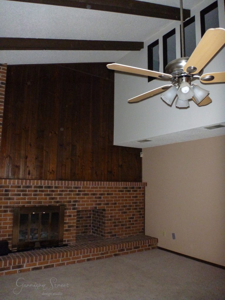 This family room, with its high ceiling, boasts a brick fireplace and a wooden accent wall that add to its casual charm. A ceiling fan with wooden blades hangs overhead. The walls are painted in neutral tones, completing the cozy atmosphere of this inviting makeover.