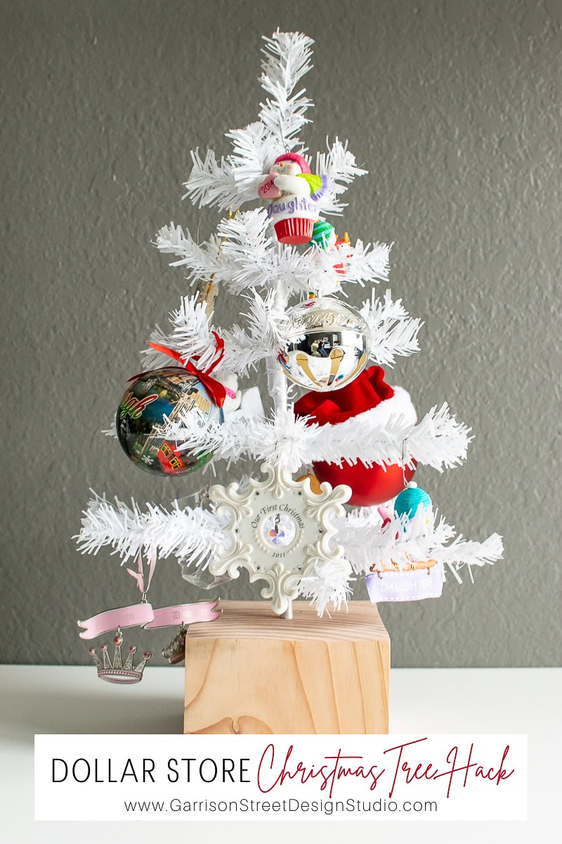 A small white Christmas tree, perfect for easy Christmas decor, is adorned with colorful ornaments like Santa, a cupcake, and a crown. It stands on a wooden block base. Text at the bottom reads: "Dollar Store Christmas Tree Hack" with a website link.
