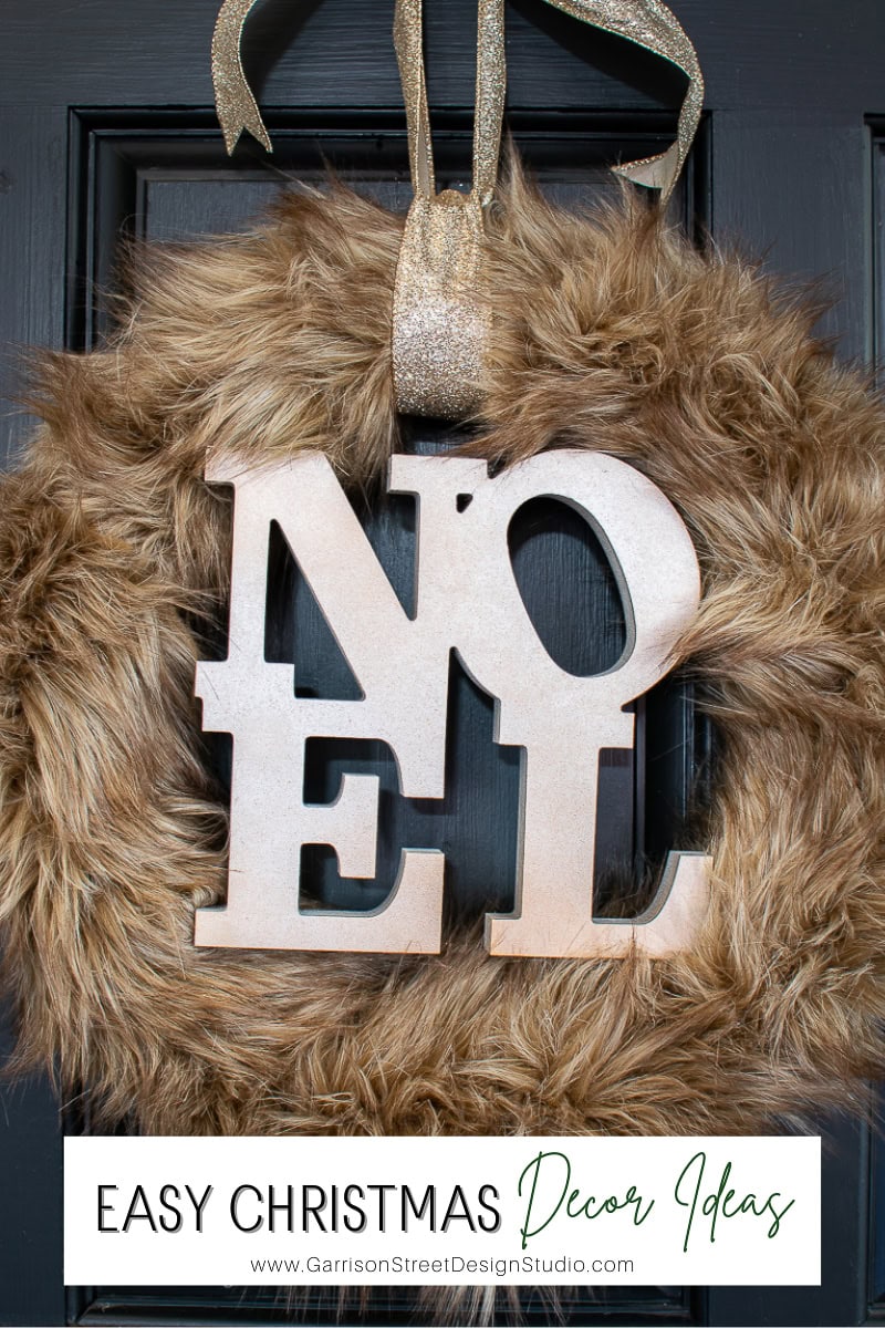 A Christmas wreath of faux fur with large wooden letters spelling "NOEL" hangs on a black door, exuding simple elegance. A gold ribbon graces the top, adding a touch of festive shine. Discover more easy Christmas decor ideas at the website link below.