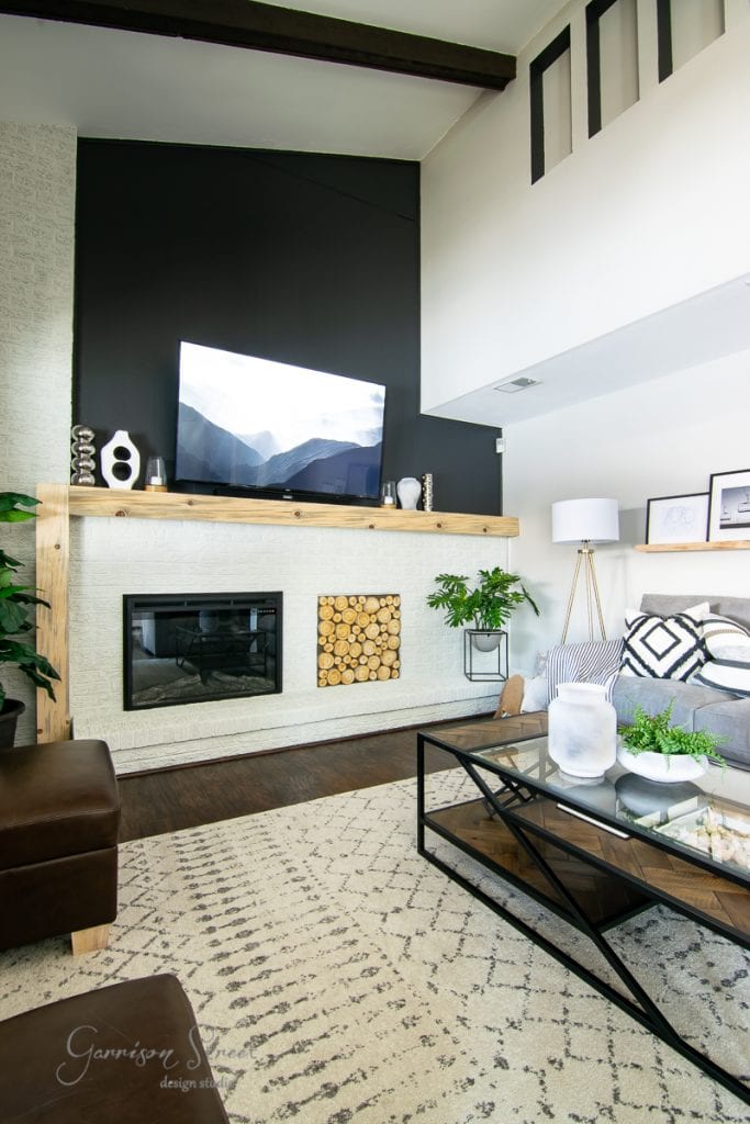 Home A modern family room featuring high ceilings, a large TV on a black accent wall, and a sleek white fireplace. The decor reveals a wooden mantel, geometric patterned rug, and glass coffee table. Indoor plants and framed pictures add greenery and style.