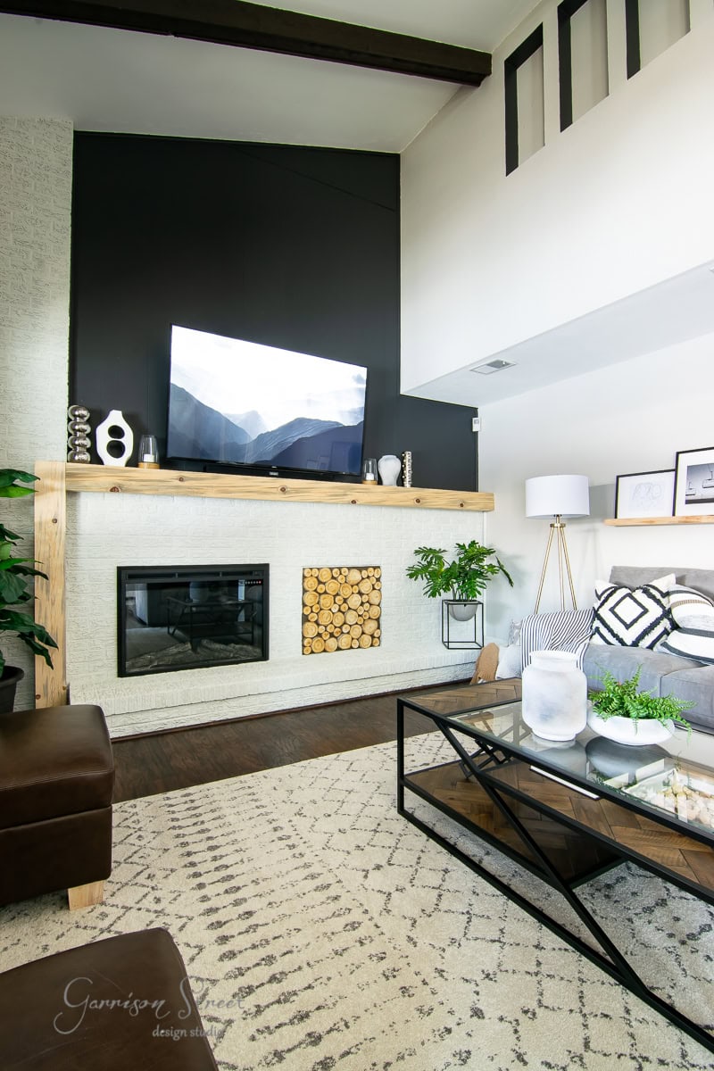A modern family room featuring high ceilings, a large TV on a black accent wall, and a sleek white fireplace. The decor reveals a wooden mantel, geometric patterned rug, and glass coffee table. Indoor plants and framed pictures add greenery and style.