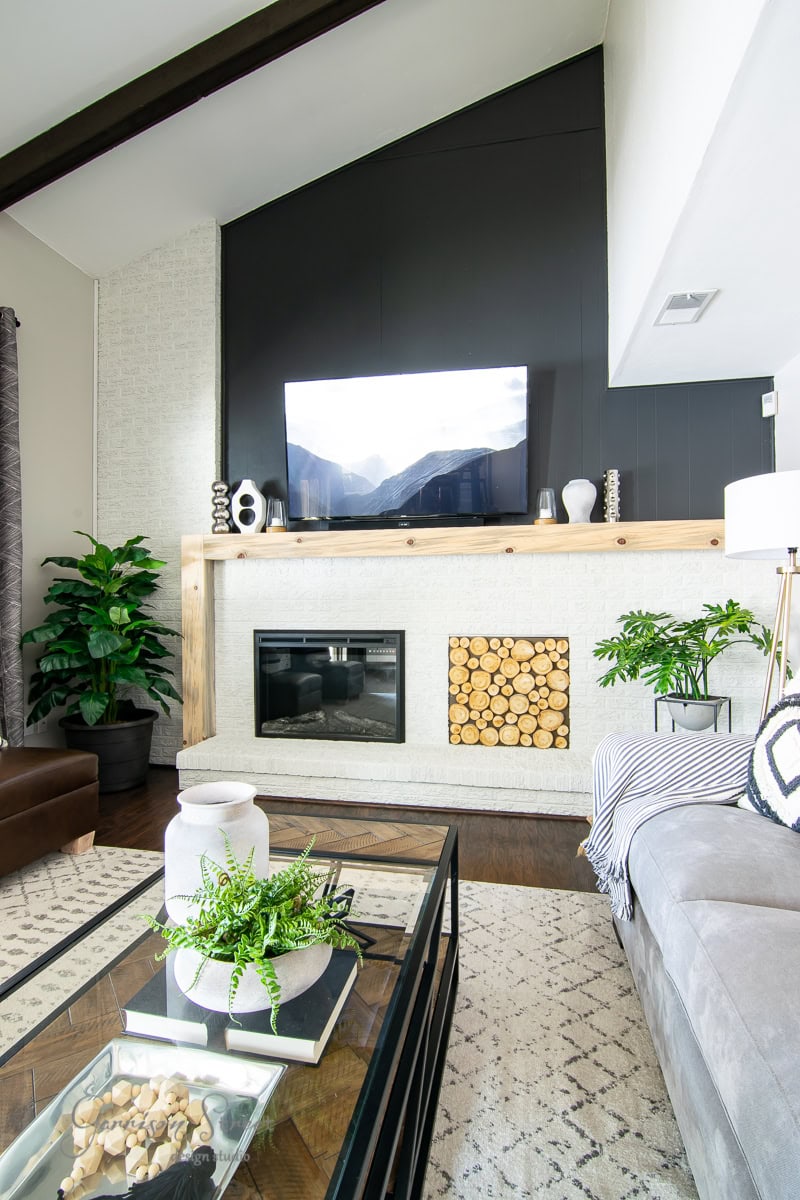 The modern family room boasts a high ceiling and a TV artfully mounted above the fireplace with log storage. A light wood mantel reveals stylish decor items. To the side, a large plant complements the coffee table, geometric rug, and sofa that complete this cozy space.