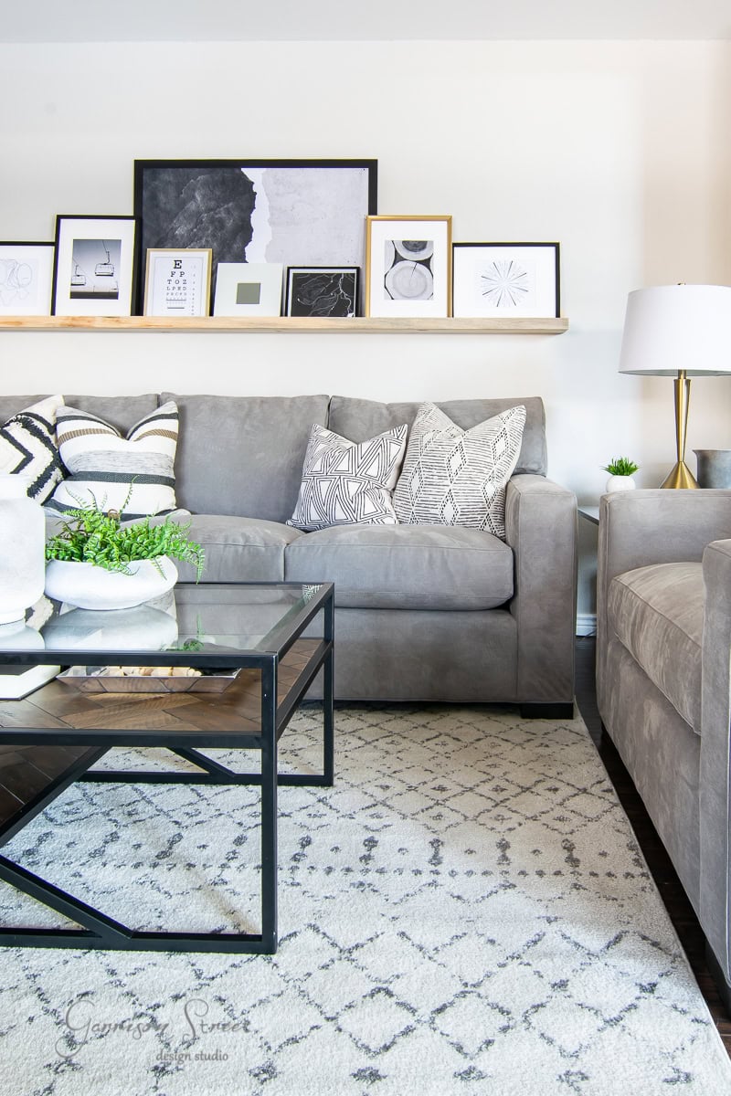 A modern family room features a gray sofa and armchair adorned with patterned cushions. A glass coffee table and beige geometric rug reveal the chic design, while framed art on a shelf above and a potted plant add greenery.