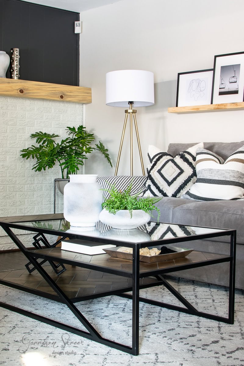 A modern family room features a gray couch with geometric patterned pillows. A minimalist black metal coffee table holds a plant and a white vase, revealing style in simplicity. A tall floor lamp stands in the corner, alongside a plant by the brick wall. Two wooden shelves display artwork.