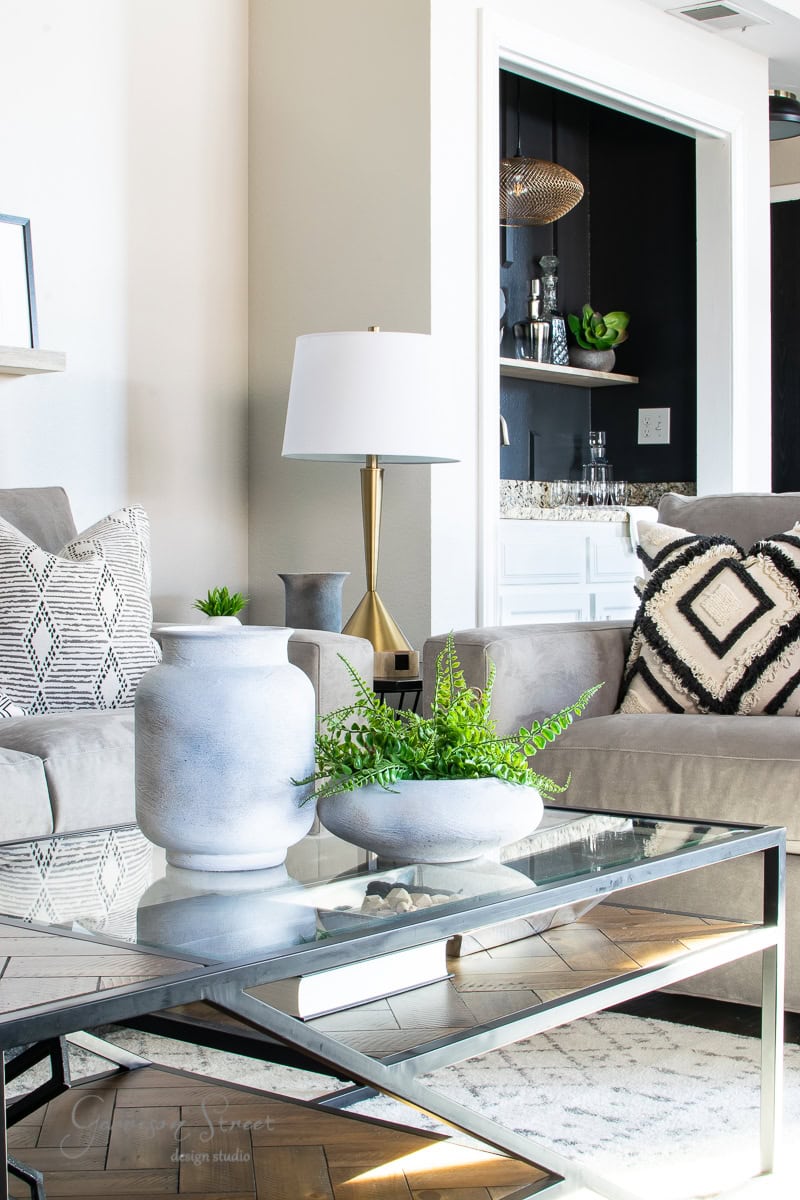 A modern family room features a glass coffee table with two white vases brimming with green plants. A gray sofa adorned with patterned pillows stands next to a sleek bar area. In the background, a white floor lamp reveals an inviting and harmonious space.