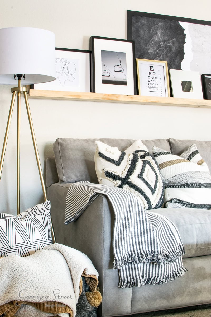 A modern, cozy family room features a gray couch adorned with patterned pillows and a striped throw blanket. Above, a wooden shelf displays framed art while a sleek lamp stands nearby, revealing a stylish and inviting atmosphere.