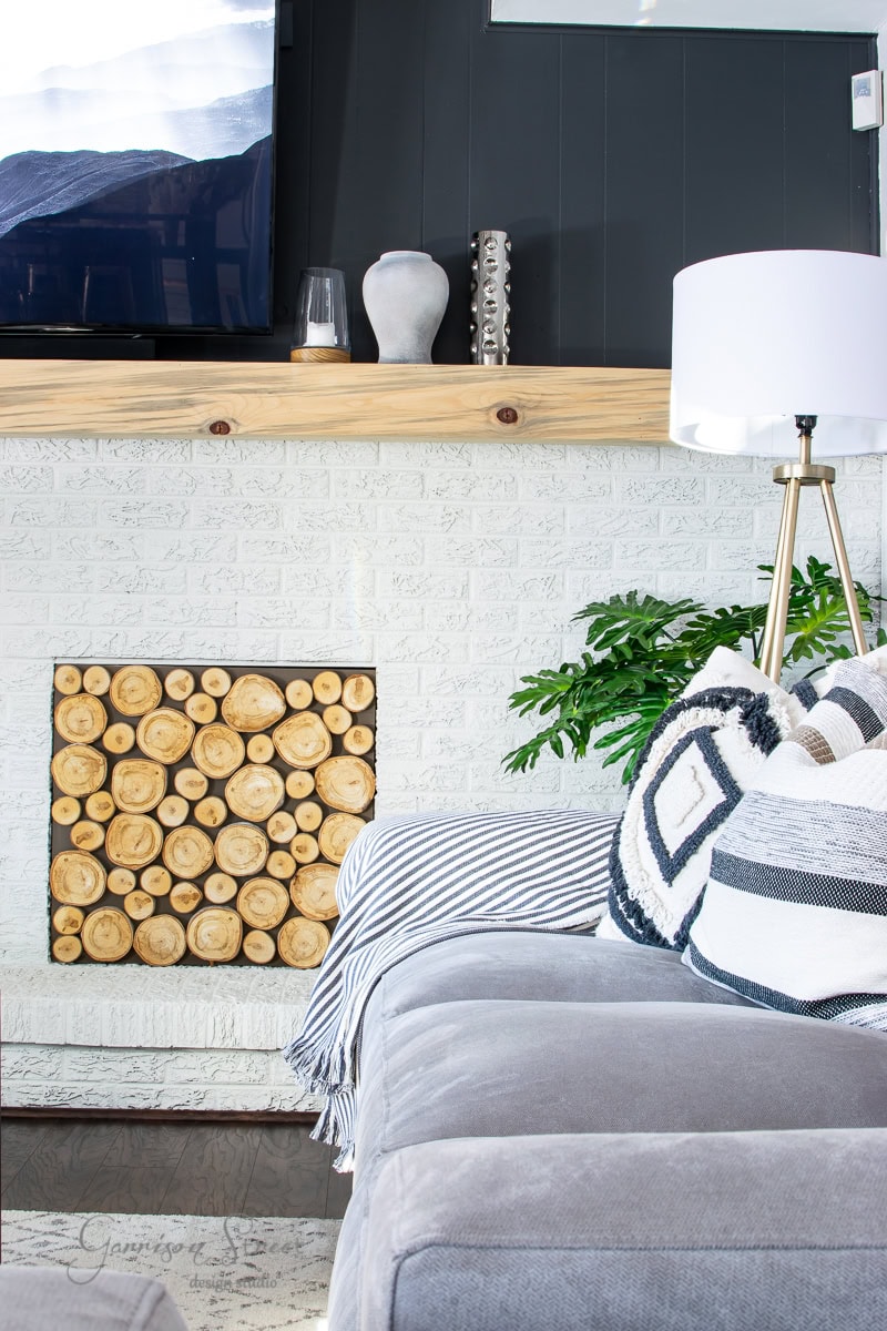 A cozy family room reveals a white brick fireplace adorned with circular wooden logs. A gray couch with patterned cushions sits nearby, while a modern floor lamp and green potted plant add warmth to the space.