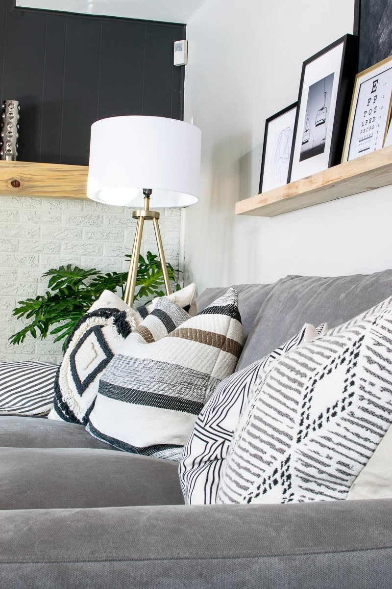 A cozy family room reveals a gray sofa adorned with patterned black and white pillows. A tall floor lamp stands beside the sofa, and framed pictures rest on a wooden shelf above. A leafy plant adds greenery to the corner, enhancing the modern aesthetic.