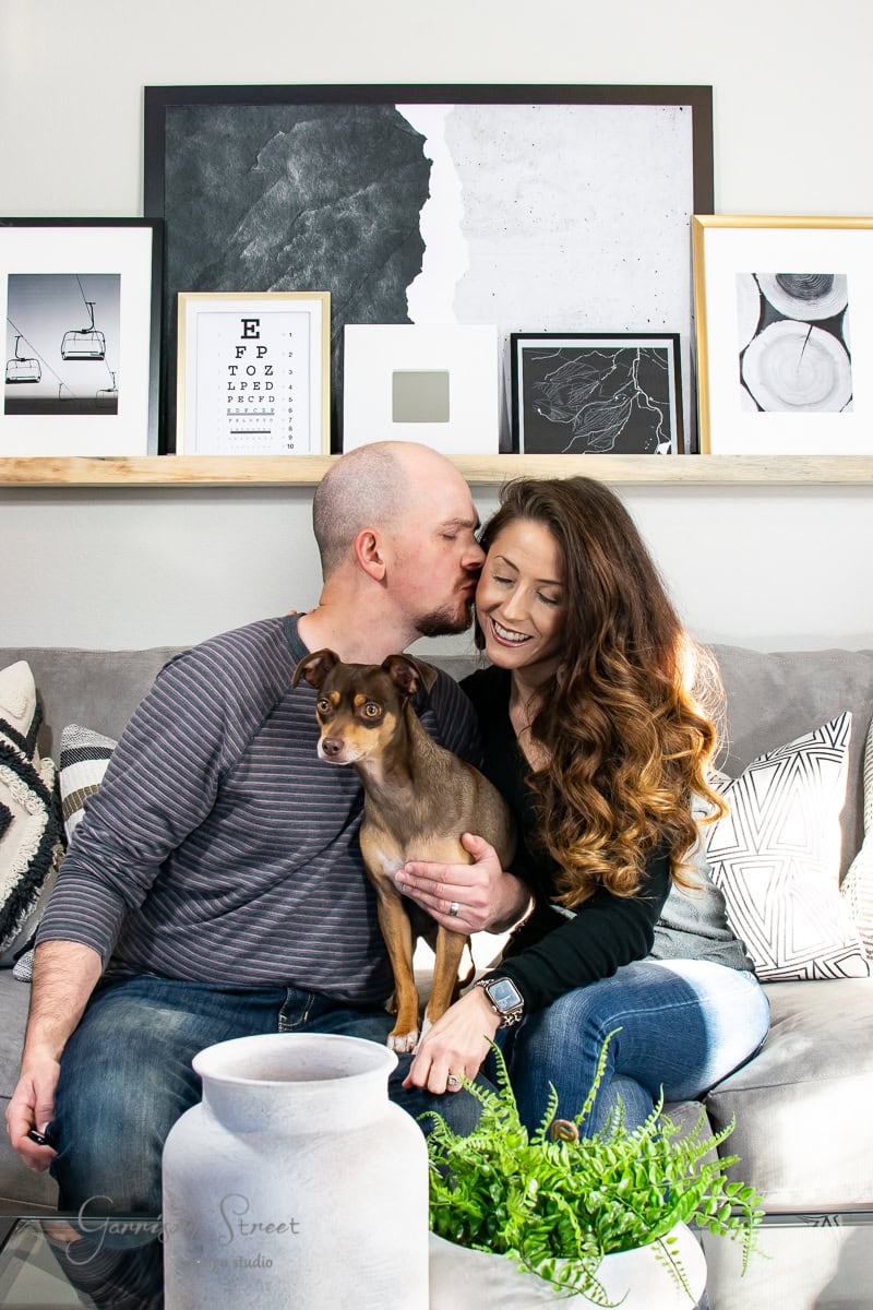 A couple sits closely on a modern couch in the family room, with the man gently kissing the woman on the forehead. She cradles a small brown dog. Behind them, framed art and photos adorn the wall, while a potted fern and decorative vase enhance the cozy atmosphere.