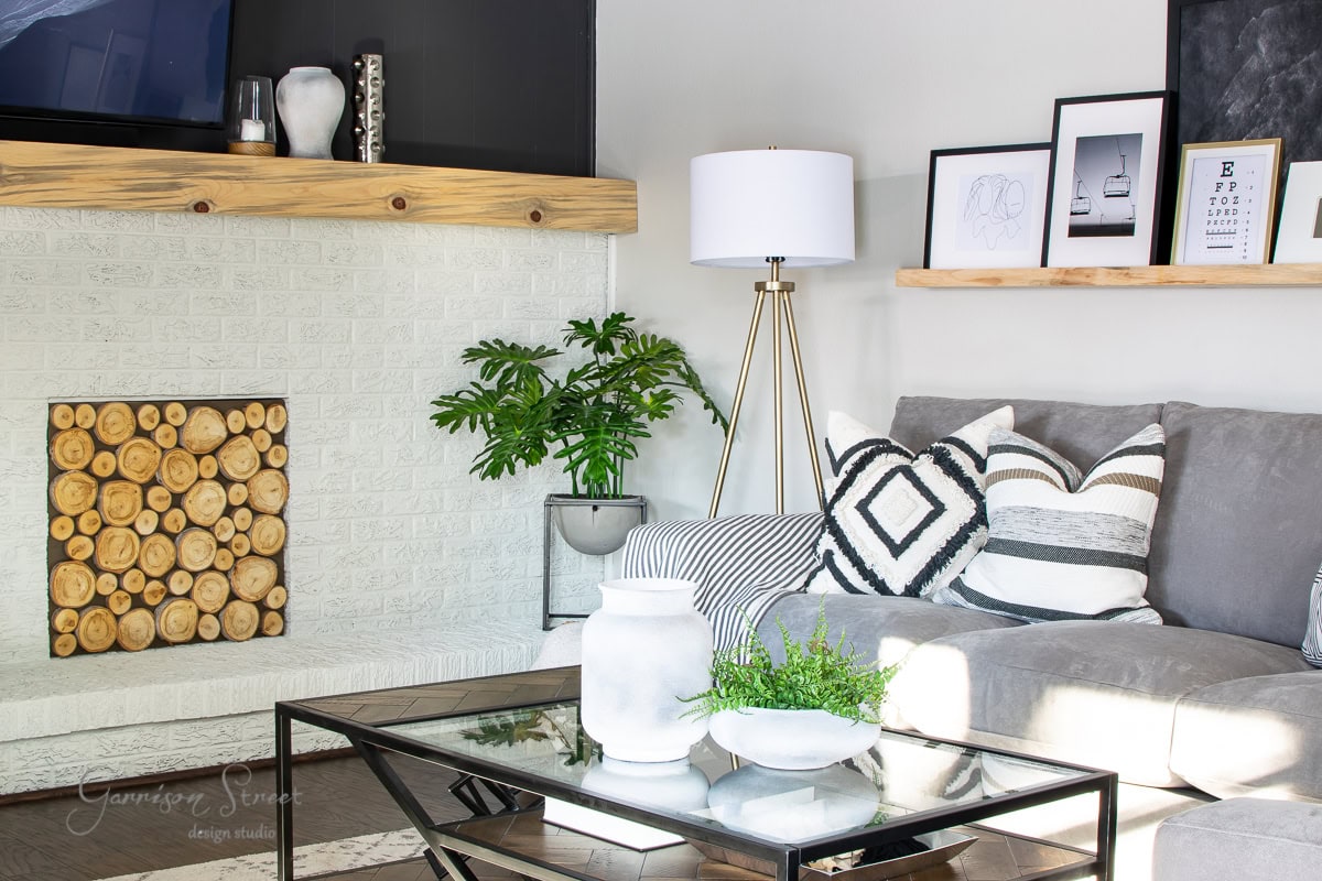 A modern family room showcases a white brick fireplace with a stack of logs, complemented by a gray sectional sofa and patterned cushions. A tripod floor lamp stands beside a glass coffee table, while framed art and a potted plant reveal the charm on a wooden shelf.