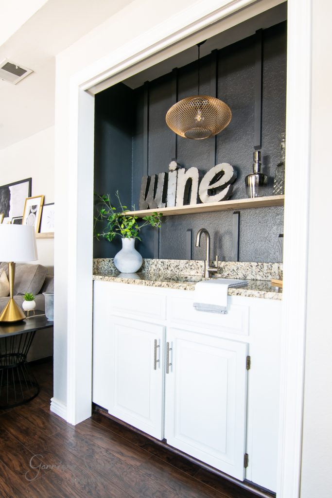 A small home bar nook with white cabinetry, a marble countertop, and a chrome faucet is revealed in the family room. A decorative "wine" sign, plants, and bottles are displayed on a shelf against a dark navy wall. A modern pendant light hangs overhead.