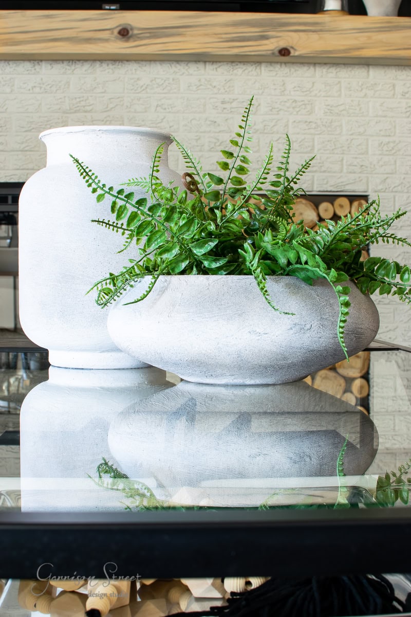 Two white ceramic pots on a reflective glass surface: a tall vase and a round bowl-shaped planter. The planter reveals lush green ferns, adding a touch of nature to the modern decor, perfectly suited for a stylish family room against a textured white brick wall backdrop.
