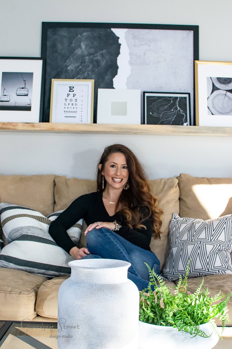 A person with long hair sits on a beige sofa, smiling in the modern family room, surrounded by decorative pillows. Behind them, a shelf displays framed art. In the foreground, a white vase and a potted fern reveal elegant touches on the table.