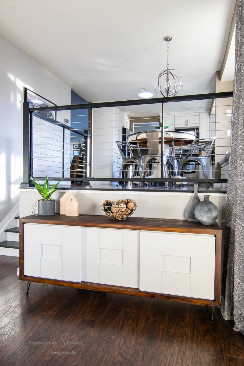 The modern interior showcases a wooden sideboard with white panel doors, perfectly blending style and function. Decor includes a potted plant, a wooden house ornament, and a metal bowl with pinecones. Beyond the reveal of the railing lies an elevated dining area with a circular table and chairs.