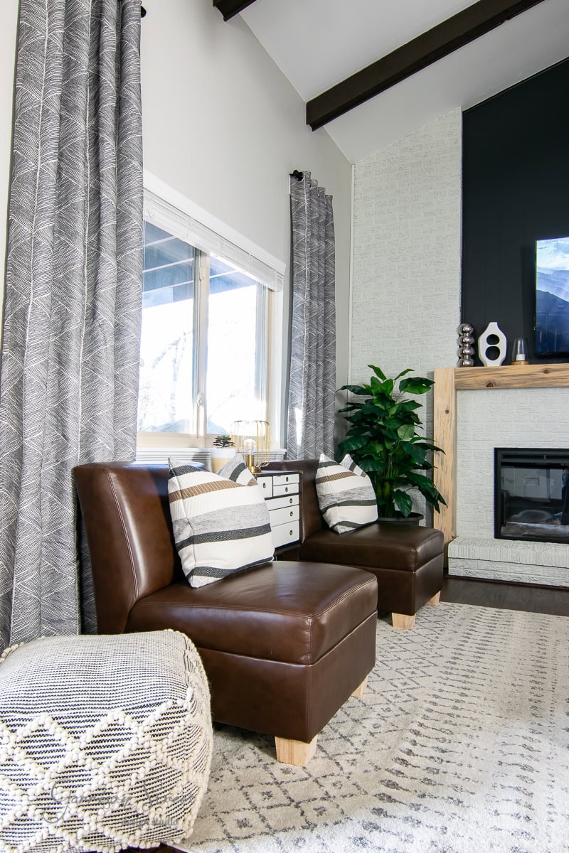 A modern family room features two brown leather chairs with striped pillows, a patterned pouf, and a woven rug. A fireplace with a wooden mantel and decorative items is on the right, while large gray curtains reveal the window view. A potted plant adds greenery.